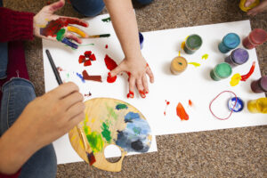une mere peint avec ses enfants, des tubes de peintures, des doigts traent des formes rouges sur un papiers